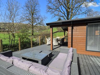 An outdoor wooden deck with wicker seating and a dining table set attached to a wooden building with a window at Jinnyspinner Lodge Beckside 37 Limefitt Park near Windermere