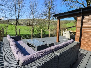An outdoor deck with a sectional sofa, glass-top coffee tables, and a dining table with chairs at Jinnyspinner Lodge in Beckside 37 Limefitt Park near Windermere