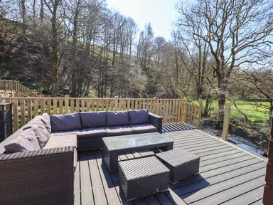 An outdoor deck with a wicker sectional sofa table and ottomans overlooking trees and a small river at Jinnyspinner Lodge Beckside 37 Limefitt Park near Windermere