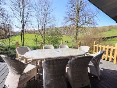 An outdoor dining area with a glass-top table and wicker chairs on a wooden deck overlooking a grassy field and trees at Jinnyspinner Lodge Beckside 37 Limefitt Park near Windermere