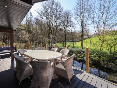 An outdoor deck with a glass table and wicker chairs overlooking a stream and trees at Jinnyspinner Lodge Beckside 37 Limefitt Park near Windermere