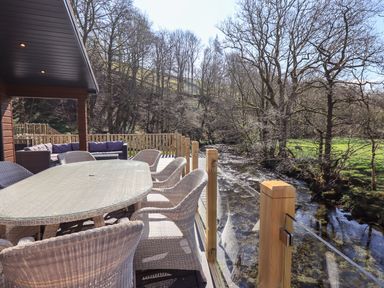 An outdoor dining area with wicker chairs and a table on a deck overlooking a river at Jinnyspinner Lodge Beckside 37 Limefitt Park near Windermere