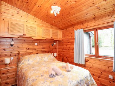 A bedroom with wooden walls a bed with a floral quilt two bedside tables and a window with blue curtains at Rowan Lodge Pinewood 16 Limefitt Park near Windermere