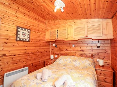 A bedroom with wooden walls and ceiling a bed with a floral bedspread and rolled towels two bedside tables with lamps and wall cabinets above the bed at Rowan Lodge Pinewood 16 Limefitt Park near Windermere