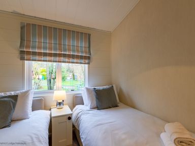 A bedroom with two single beds, a bedside table with a lamp, and a window with striped blinds at Langdale Lodge in Langdale 21 White Cross Bay near Windermere