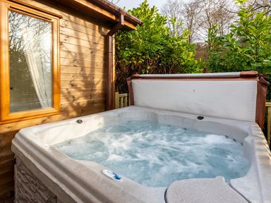 An outdoor hot tub filled with water next to a wooden house with a window and surrounded by green bushes at Langdale Lodge Langdale 21 White Cross Bay near Windermere