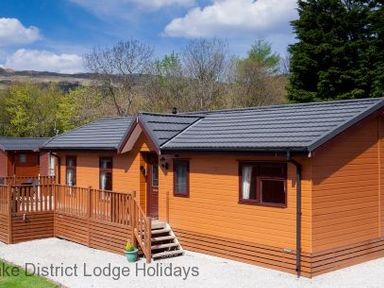 An orange wooden lodge with a grey roof and wooden deck surrounded by trees at Lake District Lodge Holidays