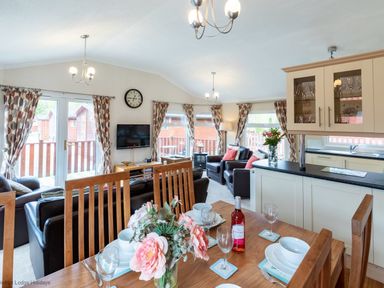 An open living and dining area with wooden dining table set with dishes and a bottle of wine a living room with sofas next to windows and blinds at Hideaway Lodge Kirkstone 14 Limefitt Park near Windermere