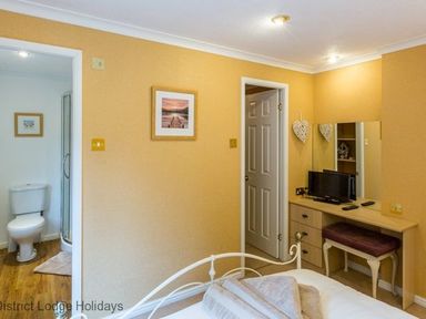 A bedroom with a bed towels on it a dressing table with a television and a stool and an open door leading to a bathroom with a toilet and shower at Hideaway Lodge in Kirkstone 14 Limefitt Park near Windermere