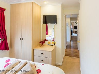 A bedroom with a bed, wooden wardrobe, chest of drawers with mirror, small TV on the wall, and red curtains at Hideaway Lodge Kirkstone 14 Limefitt Park near Windermere