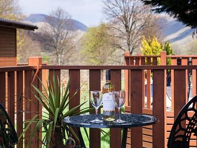 A patio table with a bottle of wine and two glasses next to a plant on a wooden balcony with trees and mountains in the background at Hideaway Lodge in Kirkstone 14 Limefitt Park near Windermere