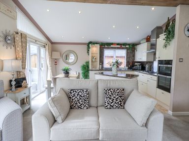 An open-plan living area with a sofa, kitchen with appliances, dining table, plants, and a wall mirror at Valley View Lodge in Wansfell 10 Limefitt Park near Windermere