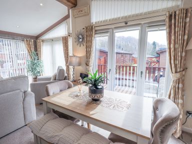 A dining table with potted plant and chairs near large windows and curtains in a living room with armchairs at Valley View Lodge Wansfell 10 Limefitt Park near Windermere
