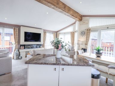 A living room with a television above a fireplace a countertop with flowers and a dining area with a bench at Valley View Lodge Wansfell 10 Limefitt Park near Windermere