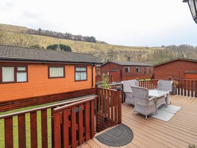 An outdoor deck with wicker chairs and a table surrounded by wooden fences and lodges in a rural hill setting at Valley View Lodge Wansfell 10 Limefitt Park near Windermere