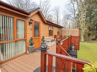 A wooden deck with benches and potted plants outside a cabin surrounded by trees and grass at Valley View Lodge Wansfell 10 Limefitt Park near Windermere