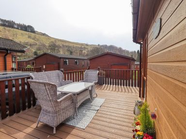 An outdoor wooden deck with wicker furniture a glass table and a floral planter on the side at Valley View Lodge Wansfell 10 Limefitt Park near Windermere