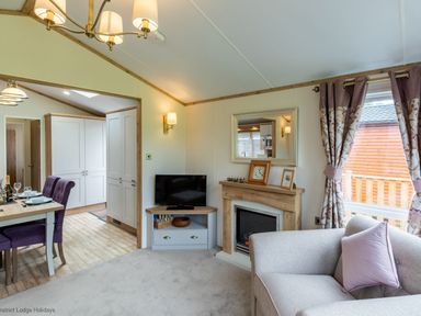 A living room with a sofa, television on a corner unit, fireplace with clock and picture frame on mantel, dining table with purple chairs, and window with curtains at Latterbarrow Lodge in Beckside 46 Limefitt Park near Windermere
