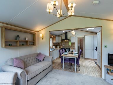 A living room with a beige sofa and purple cushions leading to a kitchen with a dining table and purple chairs at Latterbarrow Lodge in Beckside 46 Limefitt Park near Windermere