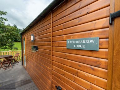 An outdoor deck with wooden chairs and table next to a wooden lodge with signs reading Latterbarrow Lodge at Beckside 46 Limefitt Park near Windermere