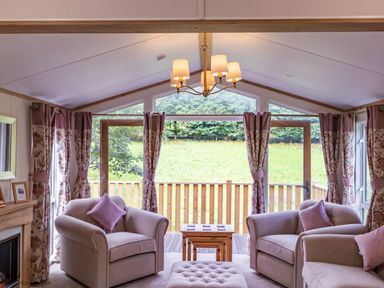 A living room with armchairs and a coffee table in front of large windows with curtains at Latterbarrow Lodge Beckside 46 Limefitt Park near Windermere