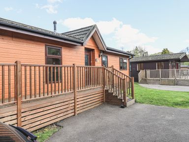 An exterior view of two wooden lodges with decks and steps on a paved area with grass at Sunny Brow Lodge Lakeside 5 White Cross Bay near Windermere