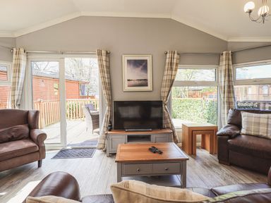 A living room with two armchairs a coffee table a television on a cabinet and windows with curtains at Sunny Brow Lodge Lakeside 5 White Cross Bay near Windermere