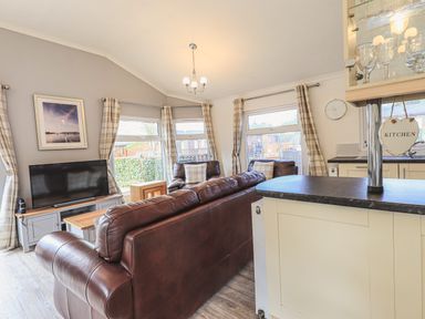 A living room with brown leather sofas a television on a wooden stand and large windows with plaid curtains at Sunny Brow Lodge in Lakeside 5 White Cross Bay near Windermere