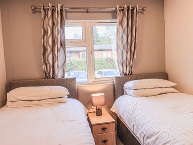 A bedroom with two single beds a bedside table with a lamp and a window with patterned curtains at Sunny Brow Lodge Lakeside 5 White Cross Bay near Windermere