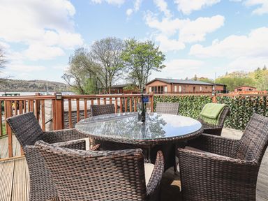 An outdoor deck with a round glass table and wicker chairs overlooking trees and wooden lodges at Sunny Brow Lodge Lakeside 5 White Cross Bay near Windermere