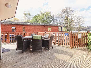An outdoor wooden deck with a round table and six wicker chairs near a wooden fence and a cabin at Sunny Brow Lodge Lakeside 5 White Cross Bay near Windermere
