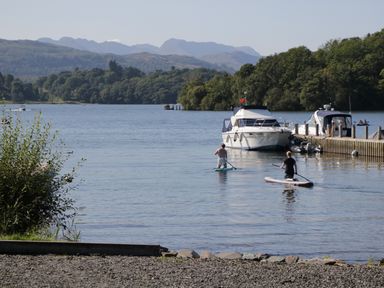 Two people paddleboarding on a lake near boats docked at a wooden pier with hills in the background at Pinehaven Lodge in Grasmere 6 White Cross Bay near Windermere