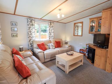 A living room with two beige sofas with red and patterned cushions a wooden coffee table a TV on a wooden cabinet and a window with patterned curtains at Pinehaven Lodge Grasmere 6 White Cross Bay near Windermere