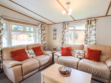 A living room with two beige leather sofas with orange and floral cushions a wooden coffee table and floral curtains at Pinehaven Lodge Grasmere 6 White Cross Bay near Windermere