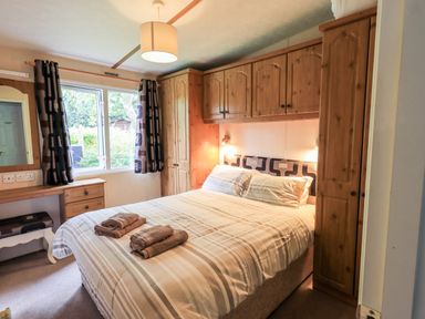 A bedroom with a double bed two brown towels built in wooden cupboards a window with dark patterned curtains and a small dressing table at Pinehaven Lodge Grasmere 6 White Cross Bay near Windermere