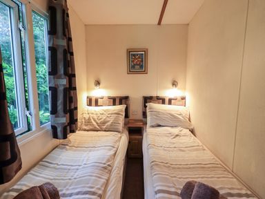 A bedroom with two single beds side by side with striped bedding a small wooden nightstand between them a window with curtains on the left wall and a framed picture on the wall Pinehaven Lodge Grasmere 6 White Cross Bay near Windermere