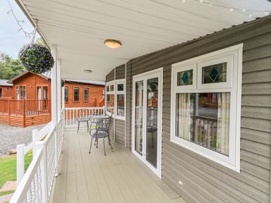 A covered wooden porch with a round metal table and chairs and hanging flower baskets at Pinehaven Lodge Grasmere 6 White Cross Bay near Windermere