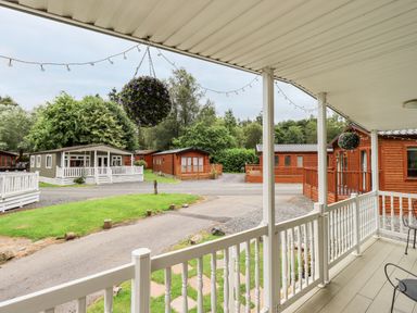 A view from a covered porch with white railings overlooking gravel paths and multiple wooden cabins surrounded by trees at Pinehaven Lodge Grasmere 6 White Cross Bay near Windermere