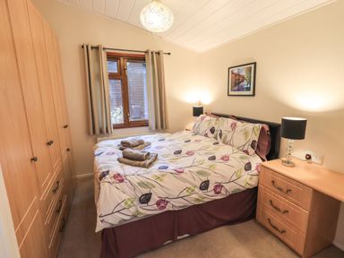 A bedroom with a bed bed with patterned bedding a window with curtains a wooden wardrobe and a wooden chest of drawers with lamps at Cedar Lodge Langdale 20 White Cross Bay near Windermere
