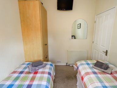 A bedroom with two single beds with checkered blankets a wooden wardrobe a wall-mounted TV a mirror a radiator and a closed door at Cedar Lodge Langdale 20 White Cross Bay near Windermere