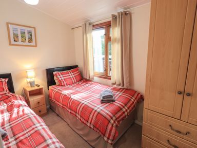 A bedroom with two single beds with red plaid bedding a wooden wardrobe a bedside table with a lamp and a window with beige curtains at Cedar Lodge Langdale 20 White Cross Bay near Windermere