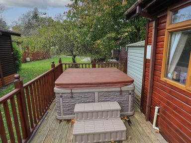 An outdoor hot tub with cover and plastic steps on a wooden deck next to a wooden cabin with a window and trees in the background at Cedar Lodge Langdale 20 White Cross Bay near Windermere