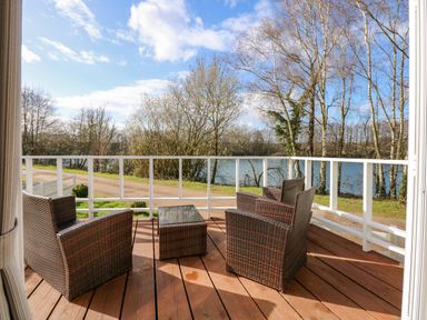 A wooden deck with three wicker chairs and a table overlooking trees and a lake at Lakeside View in Pentney near King's Lynn
