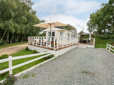 A gravel driveway leading to a mobile home with a white deck and umbrella in a wooded area at Lakeside View in Pentney near Kings Lynn