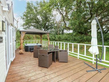 An outdoor wooden deck with a glass table and six chairs a gazebo with a hot tub and a closed umbrella at Lakeside View in Pentney near Kings Lynn