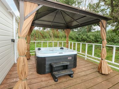 An outdoor hot tub with steps under a gazebo on a wooden deck with greenery behind at Lakeside View in Pentney near Kings Lynn