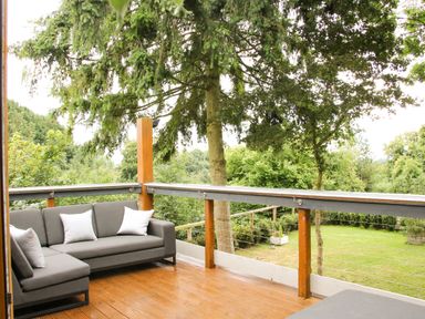 A wooden deck with gray outdoor seating and white pillows overlooking a garden with trees at Sunnyside Bircher Common near Yarpole