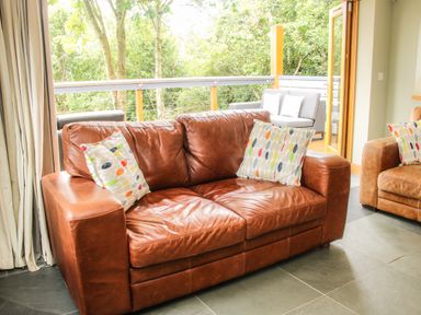 A living room with brown leather sofas and colorful pillows near an open door leading to a balcony with gray outdoor seating at Sunnyside in Bircher Common near Yarpole