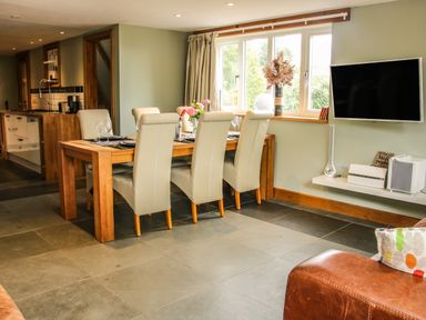 A dining area with a wooden table and six white chairs near a window and a wall-mounted TV at Sunnyside in Bircher Common near Yarpole