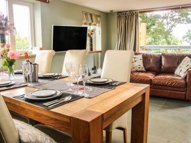 A dining table set with plates and glasses next to cream chairs and a brown leather sofa in a room with a wall-mounted television at Sunnyside in Bircher Common near Yarpole
